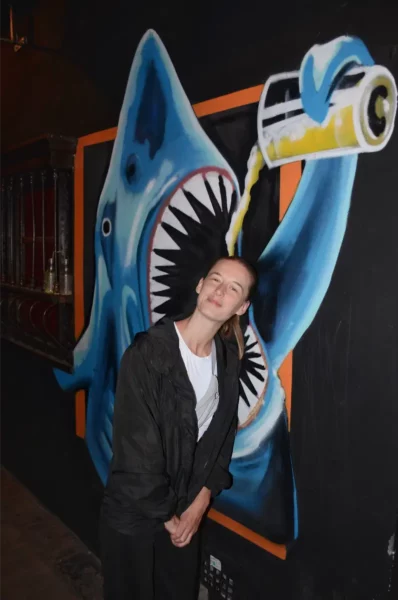 Colorful street art of a shark pouring a drink, with a smiling woman posing in front in Bogotá, Colombia.
