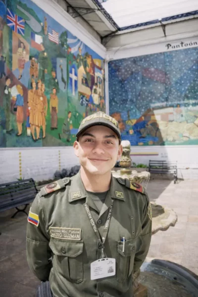 Smiling Colombian police cadet standing in the courtyard of the Police Museum of Bogotá, with colorful murals in the background