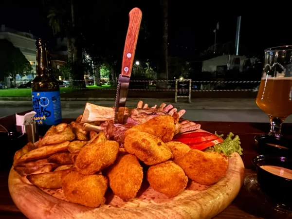 A platter of golden-brown appetizers, including croquettes and fries, accompanied by drinks, set against a nighttime Chalkis landscape.