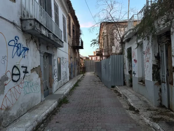 Narrow cobblestone alley lined with graffitied white buildings in Chalkis, Euboea, Greece, under a clear blue sky.