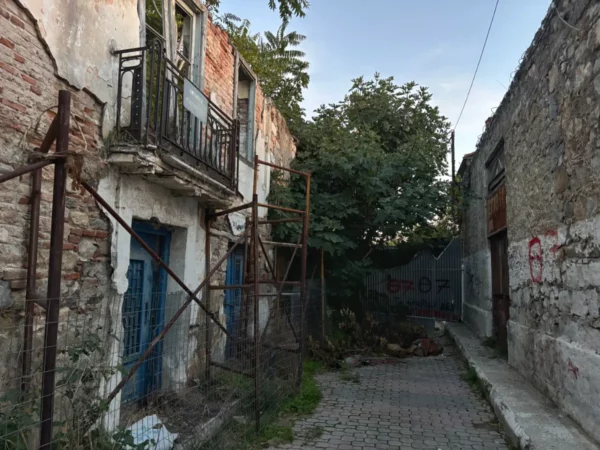 Chalkis features weathered buildings with blue windows, overgrown plants, and a cobblestone path leading through a faded urban landscape.