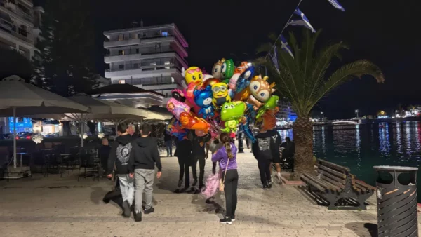 Vibrant balloons held by a group near the waterfront in Chalkis, Euboea, Greece, with palm trees and lit buildings in the background.