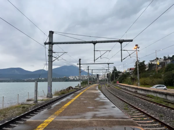 Cloudy skies loom over a quiet train station in Chalkis, Euboea, with tracks leading toward scenic mountains and the sea.