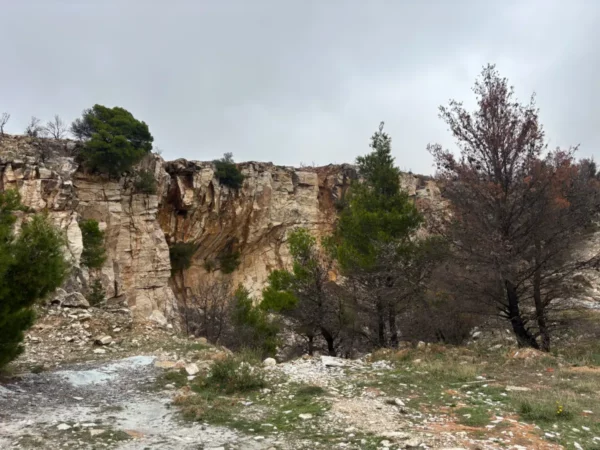 Davelis Cave features rugged cliffs and scattered pine trees on Mount Pentelicus, Attica, Greece, under a cloudy sky.