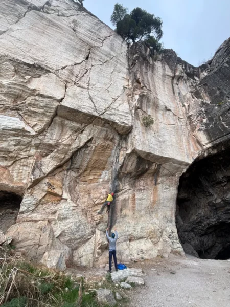 Climbers scale the rocky cliffs at Davelis Cave, surrounded by rugged textures and the greenery of Mount Pentelicus in Attica, Greece.