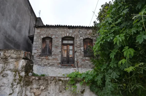 Weathered stone house adorned with wooden windows, surrounded by lush green foliage in Psachna, Euboea, Greece.