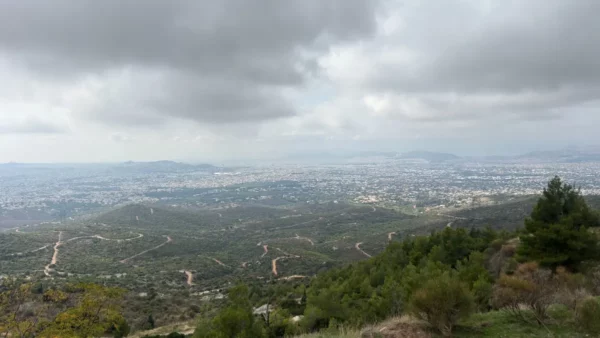 View from Mount Pentelicus in Attica, Greece, showcasing rolling green hills and a sprawling city under a dramatic cloudy sky.