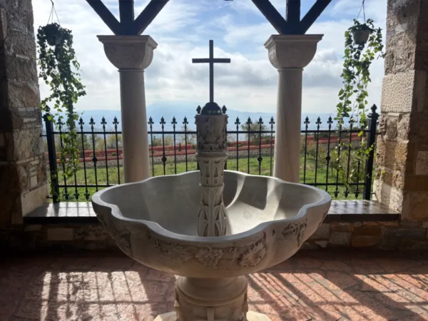 Granite fountain with a cross at Saint Panteleimon Monastery, set against lush greenery and a cloudy sky on Mount Pentelicus, Attica, Greece.