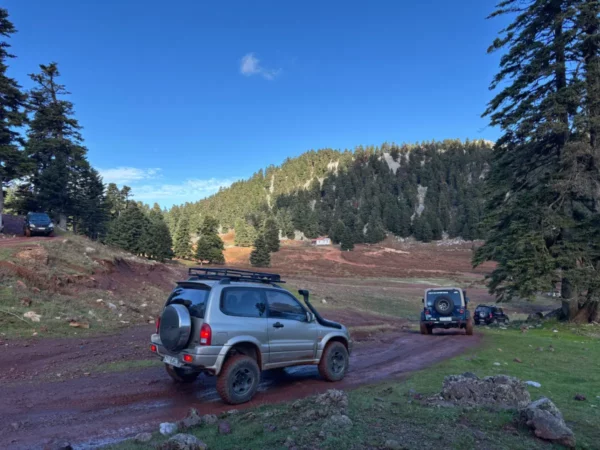 Off-road vehicles navigate a muddy track surrounded by lush greenery in Tanaida, Euboea, Greece. Bright blue skies loom above.