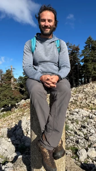Smiling traveler sits on a stone pillar, surrounded by rocky terrain and pine trees in Tanaida peak, Euboea, Greece.