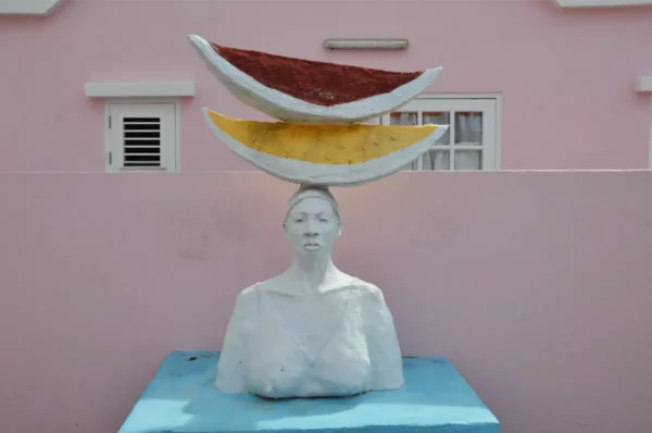 A striking statue with a woman's bust topped by colorful slices of watermelon, set against a pastel pink wall in Kaya Kaya Street Art Square, Willemstad, Curaçao.