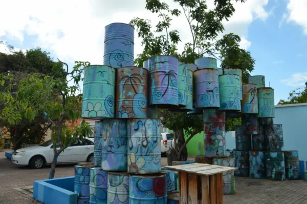 Colorful stacked barrels create an artistic installation in Kaya Kaya Street Art Square, Willemstad, Curaçao, surrounded by lush greenery and vibrant buildings.