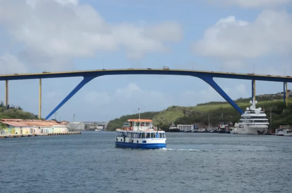 Queen Juliana bridge spans the bay in Willemstad, Curacao, with a ferry navigating the water and lush hills in the background.
