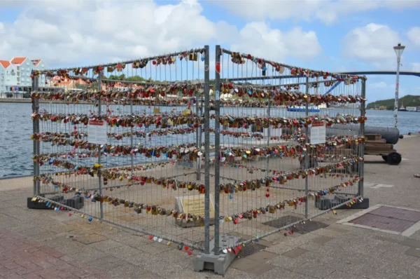 The Punda Love Heart installation: Colorful love locks adorn a metal structure by the waterfront in Willemstad, Curaçao, with vibrant buildings and the ocean in the background.