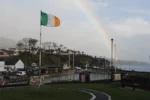 Irish flag waving near a harbor in Belfast, with a rainbow arching over lush hills and quaint buildings in the background.