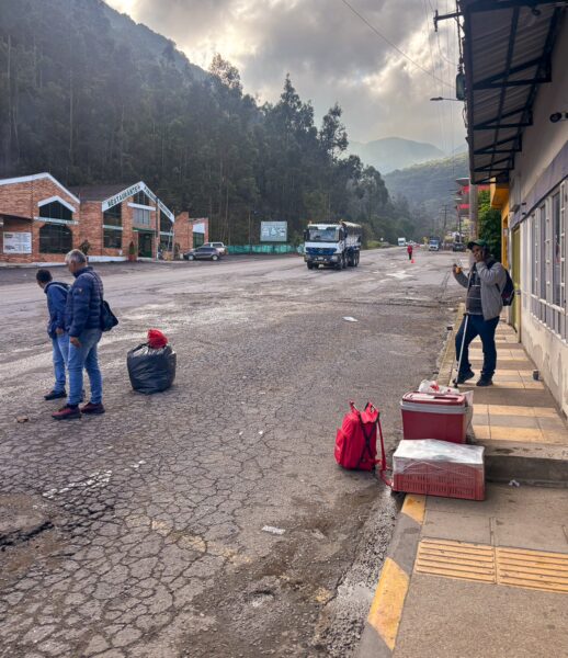 People waiting with luggage at a roadside bus stop in Arcabuco, Colombia, with mountains and misty hills in the background