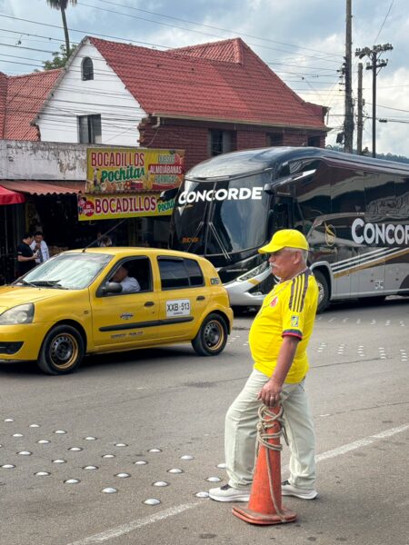 Man in a yellow shirt and cap stands by roadside traffic in Barbosa, with a bus and taxi passing a bocadillos shop.
