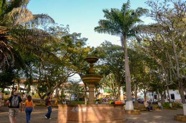 Shaded central park in Barichara with a stone fountain, tall trees, and people walking and sitting on benches