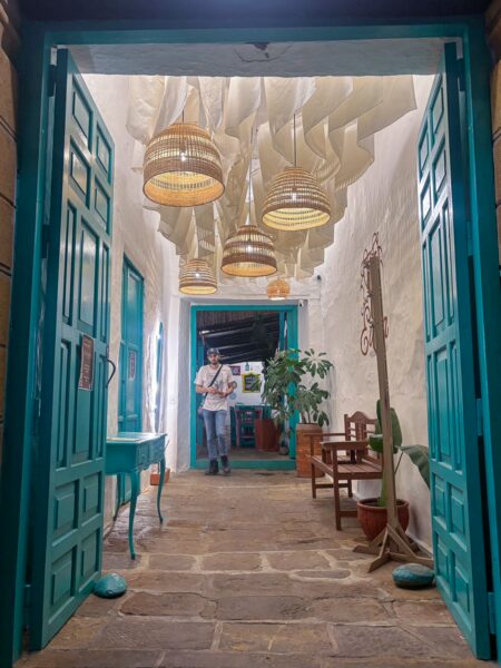 Brightly colored turquoise doors frame a charming hallway in Barichara, featuring hanging lamps and a cozy plant accent.