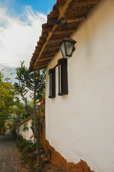 Whitewashed wall with black windows, a traditional lamp hanging, and a pathway lined with greenery in Barichara, Santander.