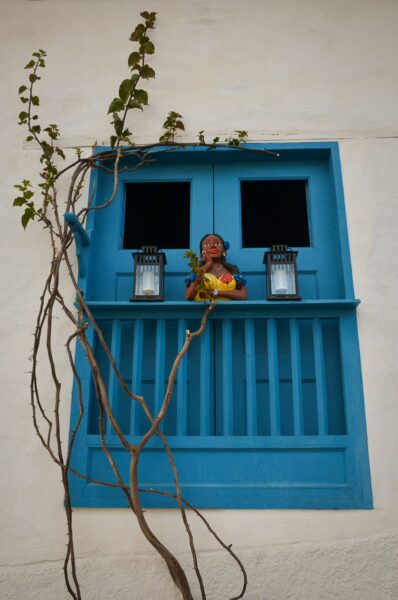 Bright blue balcony in Barichara features two lanterns and a figure holding flowers, framed by climbing vines against a white wall.
