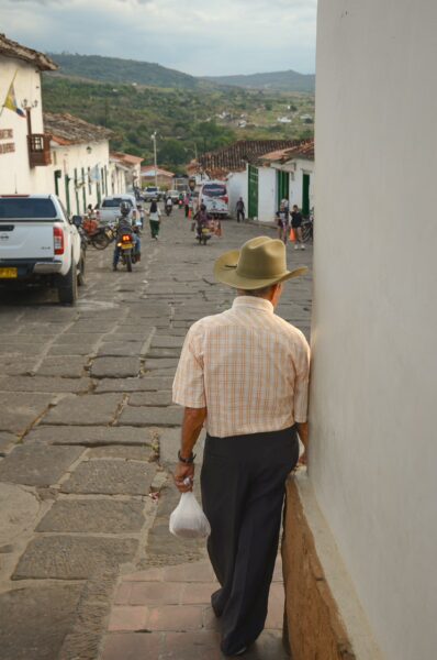 A man in a straw hat walks down a cobblestone street in Barichara, with colorful buildings and distant hills in the background.
