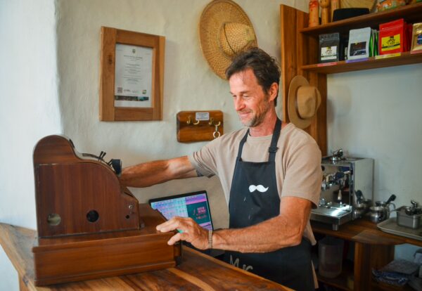 A barista operates a vintage coffee grinder in a cozy café in Barichara, Santander, surrounded by wooden shelves and coffee bags.