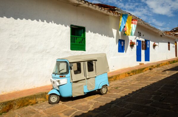 Colorful flags and a blue tuk-tuk are parked along a cobblestone street in Barichara, Colombia, against whitewashed buildings.