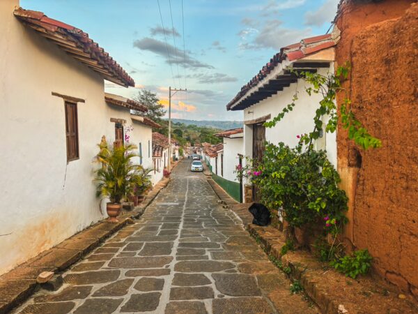 Cobblestone street in Barichara, Santander, lined with whitewashed houses and vibrant greenery under a colorful sky.