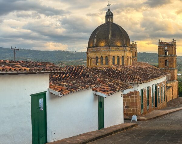 Traditional whitewashed houses with green doors and a domed church stand out against a dramatic sky in Barichara, Santander, Colombia.