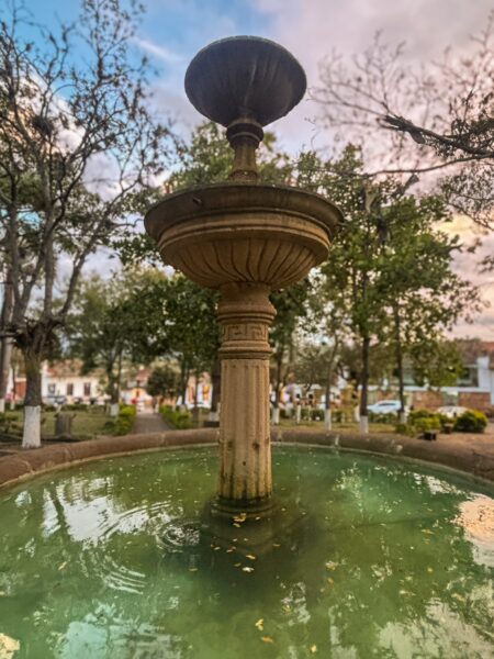 Historic fountain surrounded by lush greenery in Barichara, Santander, Colombia, reflecting a vibrant sunset sky.
