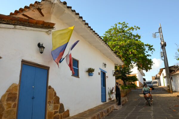 Brightly colored flags adorn a whitewashed house with a blue door in Barichara, Colombia, while a cyclist passes by.