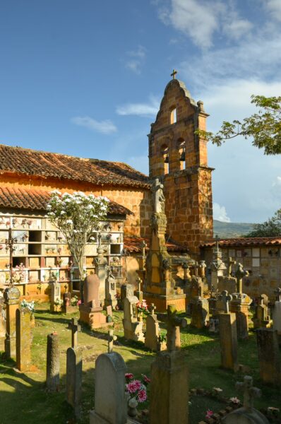A serene cemetery in Barichara, featuring weathered tombstones, vibrant greenery, and a historic church tower against a blue sky.