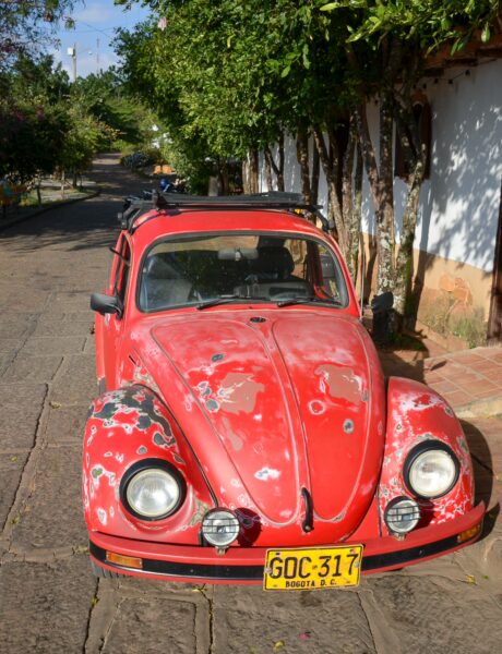 Classic red Volkswagen Beetle parked on a cobblestone street in Barichara, Santander, surrounded by green trees and rustic buildings.