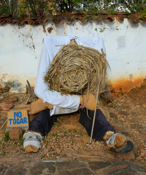 A scarecrow dressed in white with a straw hat sits against a wall in Barichara, surrounded by rustic stones and a "Do Not Touch" sign.