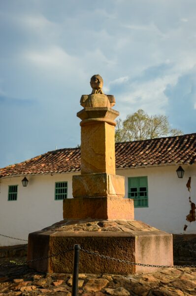 Small Simón Bolívar monument on a stone pedestal, with a white colonial building and tiled roof behind it in Barichara