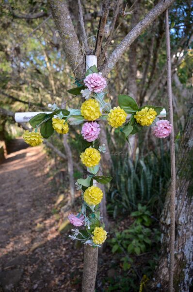 A decorated cross adorned with pink and yellow flowers, set against a lush green background in Barichara, Santander, Colombia.