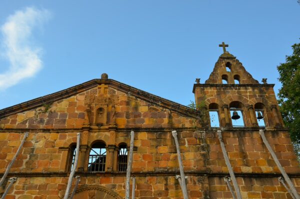 The rustic stone façade of a church in Barichara, Santander, features arched windows and a tall bell tower against a blue sky.