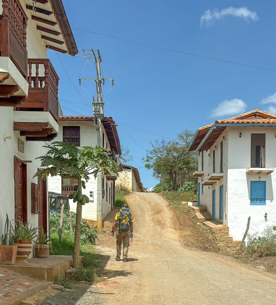 A traveler strolls along a sunlit dirt road lined with charming white houses and colorful doors in Barichara, Santander.