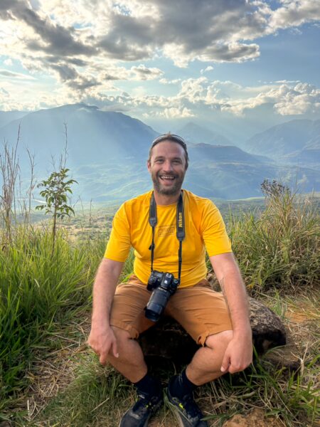 A smiling man in a yellow shirt sits on a log with a camera, surrounded by lush green hills of Barichara, Santander, under a cloudy sky.
