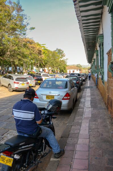 A motorcyclist rests beside a line of parked cars on a cobblestone street in Barichara, surrounded by lush greenery.