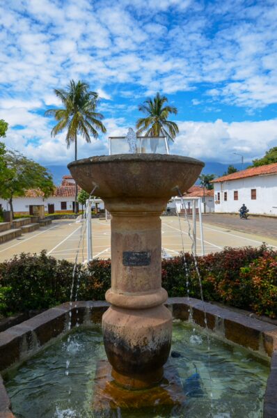 Stone fountain in Guane’s main square, with a basketball court, white buildings, and palm trees under a cloudy blue sky