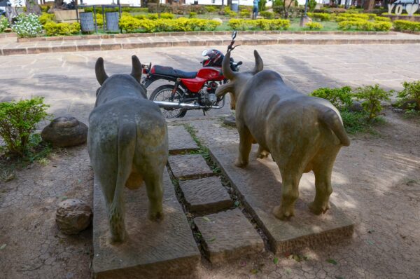 Stone sculptures of two bulls gaze towards a red motorcycle in a park in Guane, Santander, Colombia, surrounded by greenery.