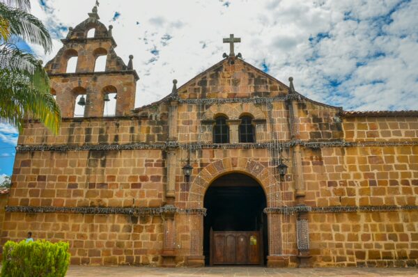 Stone church facade in Guane, Santander, with an arched entrance, bell tower, and cross against a cloudy sky