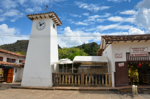 White clock tower stands alongside quaint buildings in Guane, Santander, Colombia, beneath a bright blue sky. A dog rests nearby.