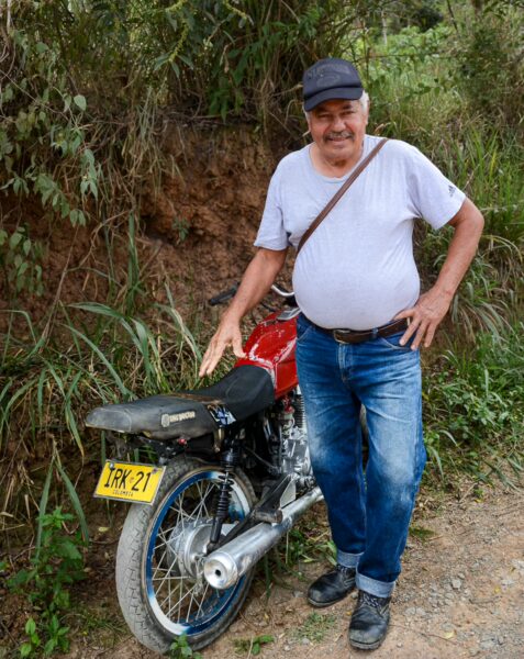 A man stands beside a red motorcycle on a rural road in Guane, Santander, Colombia, surrounded by lush greenery and hills.