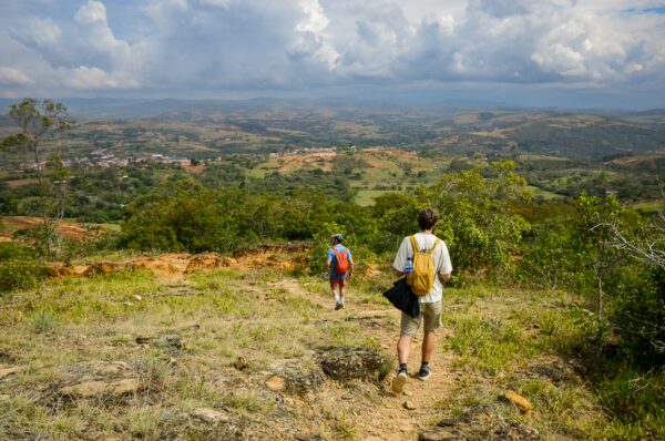 Hikers descend a trail through lush greenery in Guane, Santander, Colombia, with rolling hills under a cloudy sky.