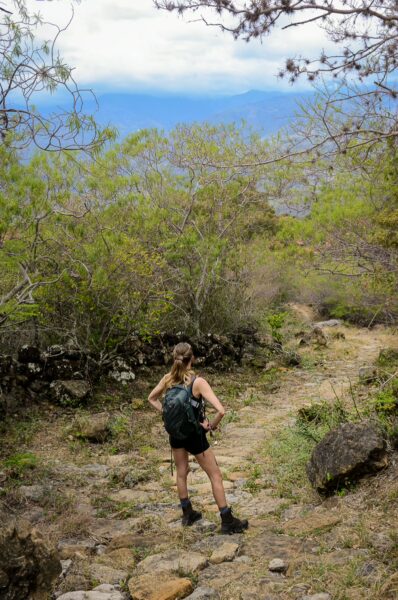 A hiker pauses on a rocky trail surrounded by green foliage in Guane, Santander, Colombia, with mountains in the distance.