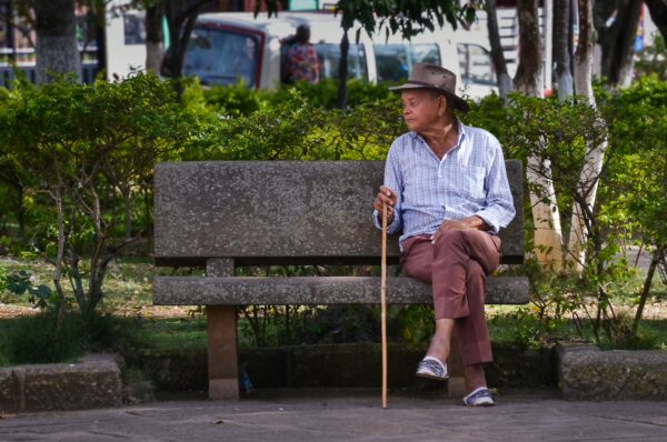 Man in a hat sits on a park bench with a walking stick in Villanueva, Santander