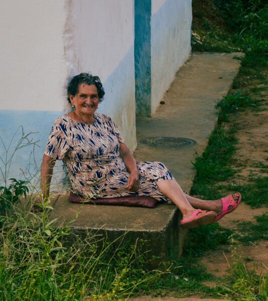 A woman in a patterned dress sits on a stone step by a wall painted in light blue, surrounded by lush greenery in Villanueva, Santander.